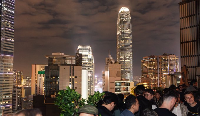 A nighttime view of a city skyline featuring tall, illuminated skyscrapers under a cloudy sky, with a crowd of people gathered in the foreground, suggesting a rooftop bar or event.