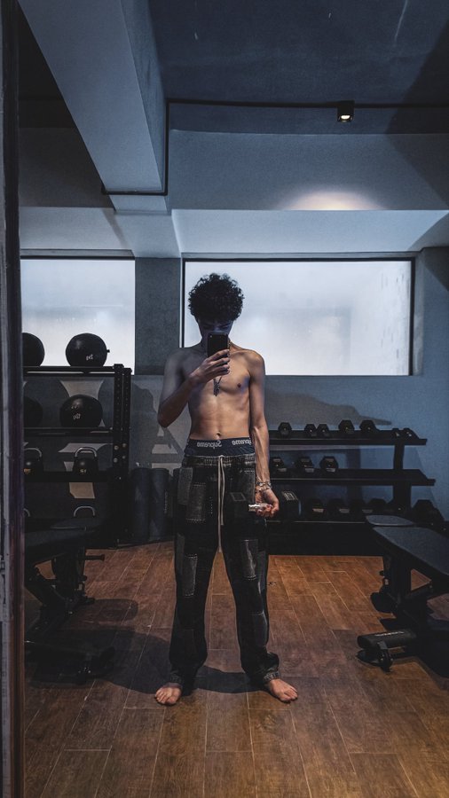 A shirtless man with curly hair takes a mirror selfie in a gym, holding a phone while wearing dark pants and standing barefoot on a wooden floor surrounded by weights and medicine balls.