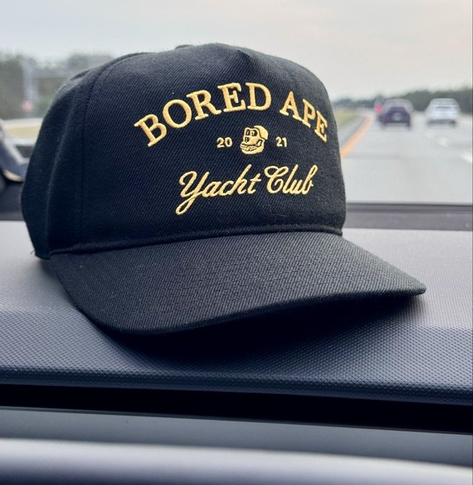 A black snapback cap with gold embroidered "Bored Ape Yacht Club 2021" text sits on a car dashboard, with the post's #BAYC hashtag and "Don't miss the boat" tagline referencing the famous NFT collection.