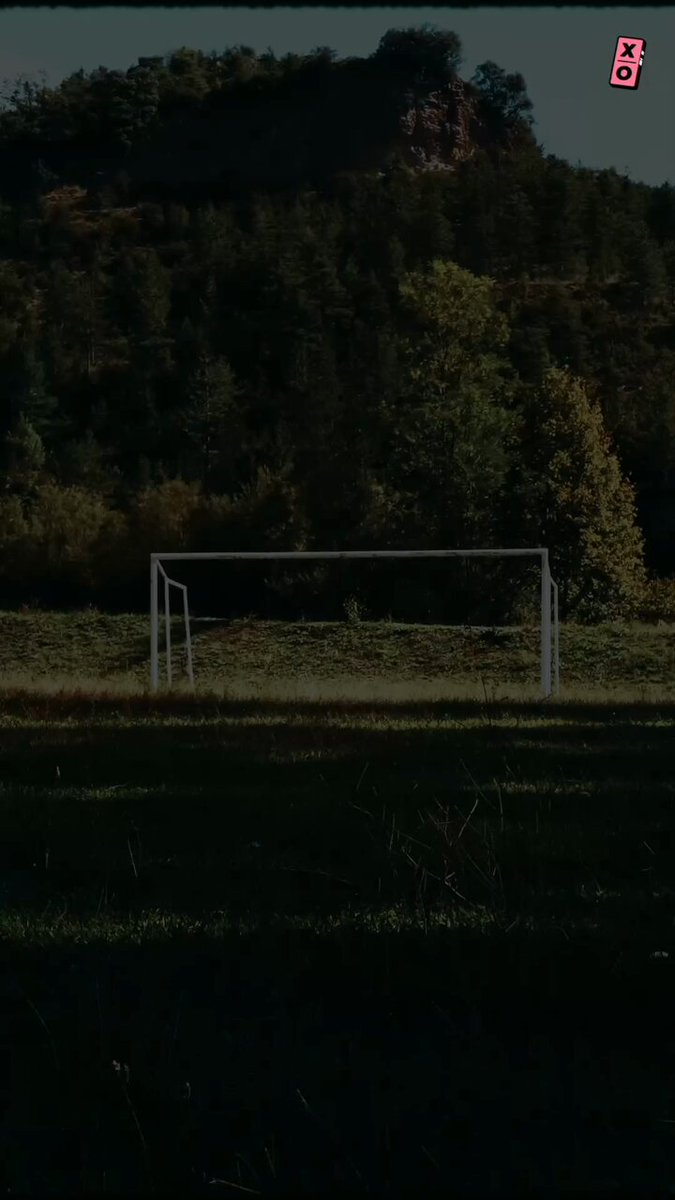 The image displays a soccer field with a white goalpost sitting within an open grassy area under a tree line. There are no people visible in the scene, suggesting it's either during off-season or has been captured at night when there are fewer people around.
