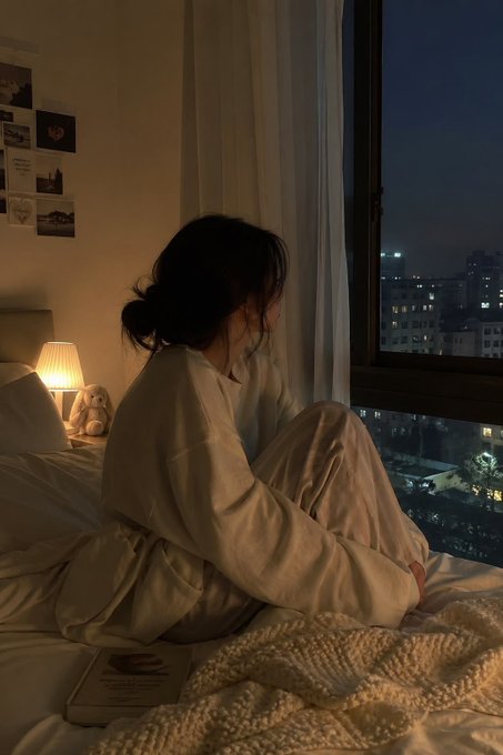 A young woman sits on her bed in cozy pajamas, gazing out a window at a dimly lit city skyline at night, with a warm lamp and Polaroid-style photos creating an intimate atmosphere that mirrors the late-night, winding-down vibe of the post's "Goodnight" message.