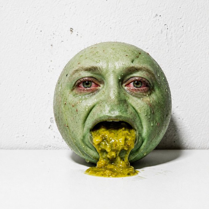 A surreal sculpture resembling a green, round human face with an open mouth vomiting yellow-green liquid, displayed on a white surface against a textured white wall.