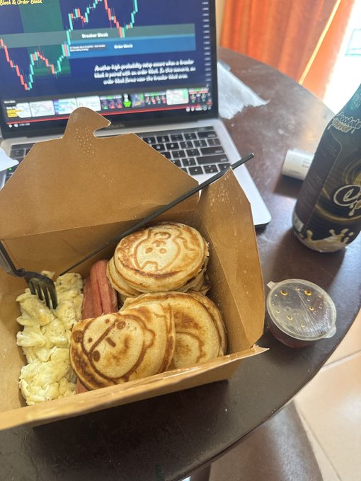 A cardboard takeout box holds smiley-face pancakes, scrambled eggs, and a sausage link, paired with a small syrup cup, while a laptop displaying a "Boulder Brush" stock chart and a Chocoddle drink sit in the background—fitting the "G to the M" hype as the trader fuels up before making market moves.