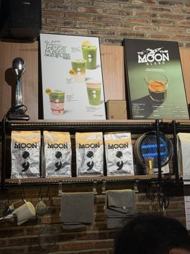 The image shows a promotional display for coffee brand "Moon Coffee Co." featuring an Ecuador Machamba poster alongside bags of coffee beans and merchandise, reinforcing the post's mocking by labeling the seller as "stinky" while showcasing Moon Coffee's specialty offerings. The brand's logo is prominently displayed on both the menu boards and packaging, with a spotlight illuminating the display against the brick wall backdrop.