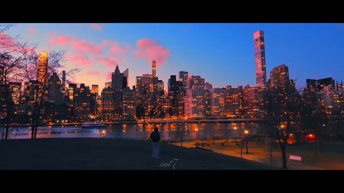 The image shows a city skyline at sunset, with a person standing near the water's edge of a park or riverfront area. The sky has beautiful pink hues, creating a serene atmosphere for the scene. In the background, there are tall buildings that add depth and perspective to the view. The photo appears to be taken from a distance, capturing both the cityscape and the person enjoying their time by the water.