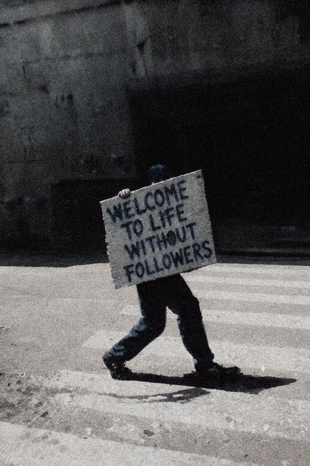 A person walks across a crosswalk holding a large sign that reads 'WELCOME TO LIFE WITHOUT FOLLOWERS'.