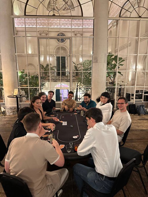 A group of around nine friends gathers around a poker table in an elegant, vaulted-ceiling venue, enjoying a lively card game with drinks in hand. The image captures the social atmosphere referenced in the post, highlighting the camaraderie of the Superteam UK gathering with special guest vibhu as they enthusiastically discuss Solana ($SOL) and other crypto investments.