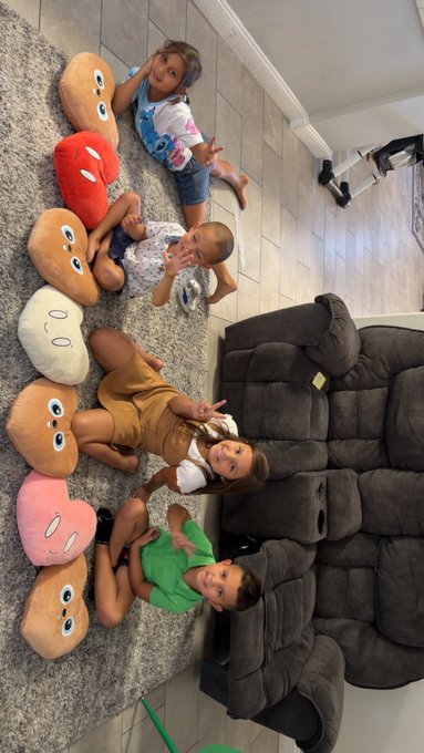 Four children sit on a carpeted floor next to a row of six smiling emoji-style plush pillows in various colors (tan, red, white, brown, pink, and tan), making peace signs and posing for the camera during what appears to be a fun NFT collab showcase.