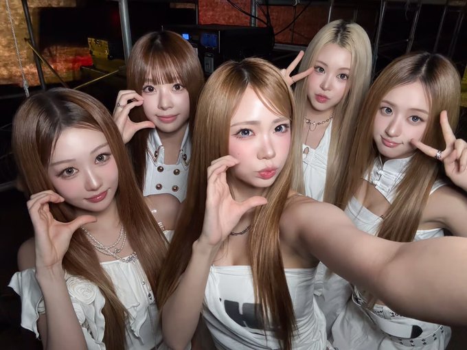 Five young women with long hair pose together for a selfie, wearing white outfits and making hand gestures near their faces.