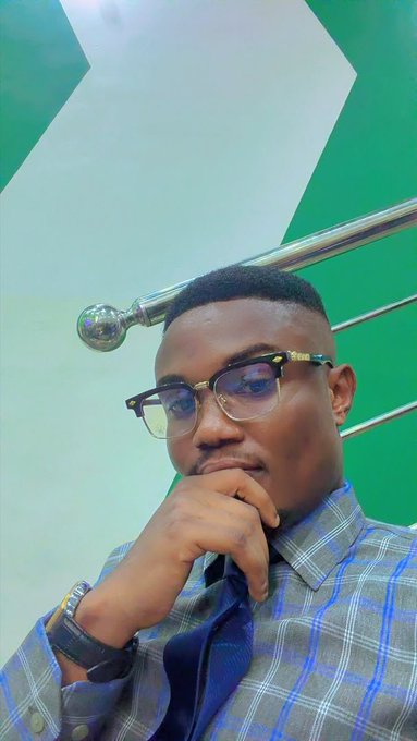 A selfie of a young man wearing glasses and a grey plaid shirt, posing with his hand near his chin in front of a green wall with a metallic railing.