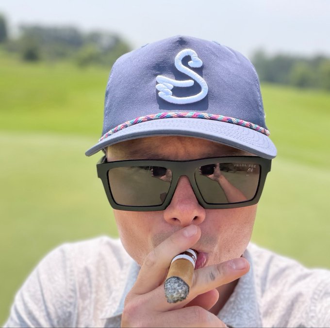 A selfie of a man wearing a dark baseball cap with a stylized 'S' logo (Seattle Kraken) and dark sunglasses, smoking a cigar on what appears to be a golf course.