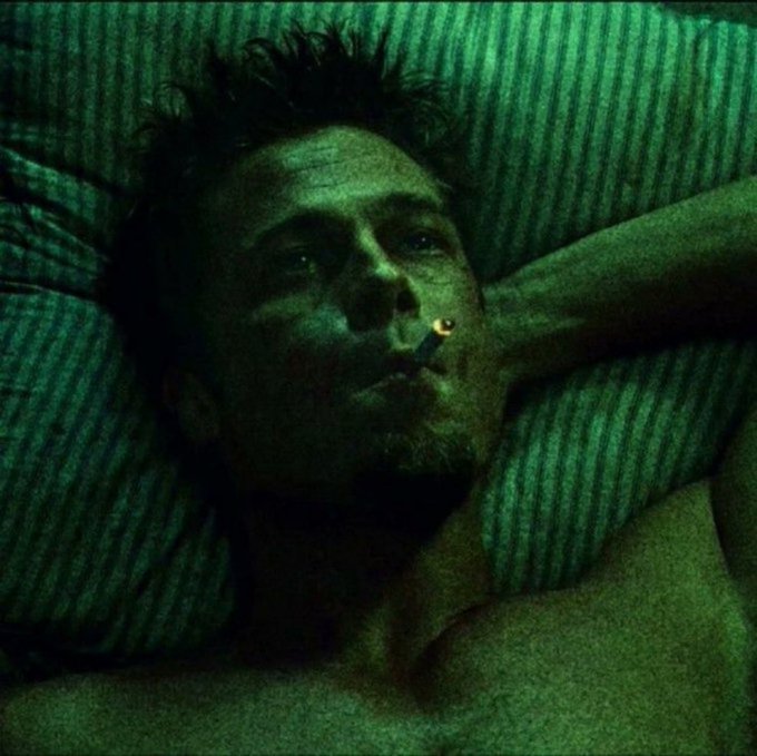 A moody, dimly-lit close-up of a young man with messy hair and a goatee lying on striped bedding, smoking a cigarette, with a green-tinted cinematic atmosphere reminiscent of the film "Requiem for a Dream."