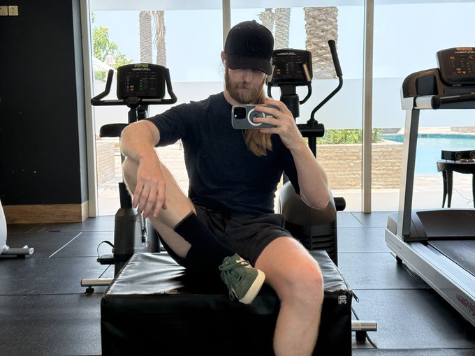 A bearded man in a black cap and athletic wear takes a mirror selfie while sitting casually on gym equipment in a modern home gym, with palm trees and a pool visible through the windows behind him. The post pairs this relaxed, confident pose with a tough-talking caption encouraging perseverance and mocking those who shy away from challenges, using the fitness setting as a metaphor for mental grit.