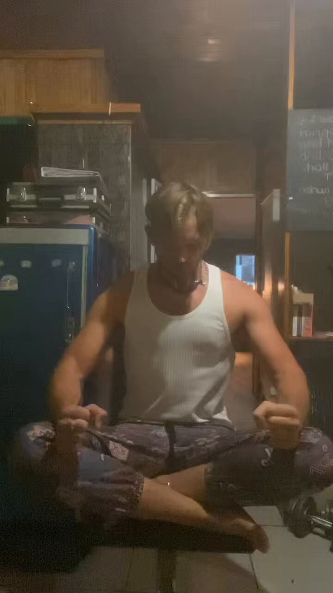 The image shows a man sitting cross-legged with his hands folded, wearing a white tank top and blue pants. He appears to be meditating or practicing yoga indoors. The room has wooden cabinets and a refrigerator visible in the background. There is also a book on one of the cabinets, suggesting that this space could serve as a personal sanctuary for relaxation and self-reflection.