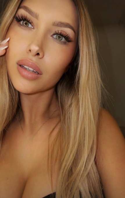Close-up portrait of a blonde woman with green eyes, long lashes, and glossy nude lipstick, wearing a dark top.