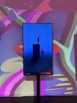 An underwater sculpture installation featuring figures standing on pedestals is displayed on a vertical digital screen mounted on a stand.