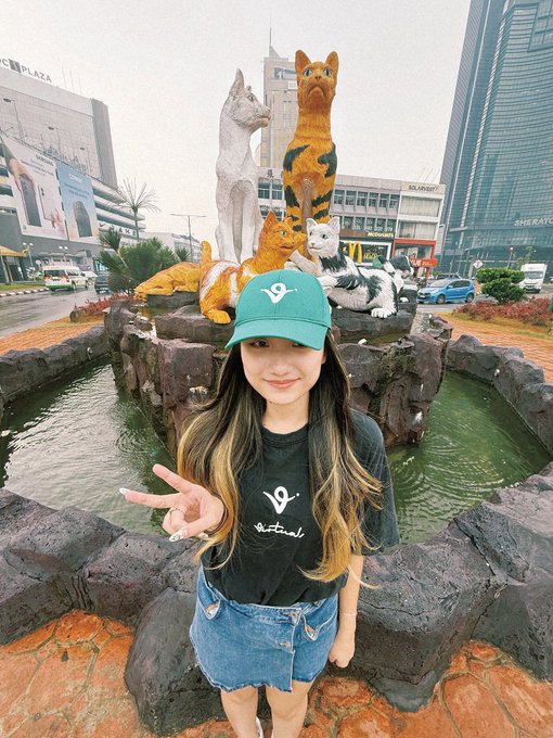 A young woman wearing a green Virtuals cap and t-shirt poses with a peace sign in front of the famous cat statues at Jeju Island's Cat Park.