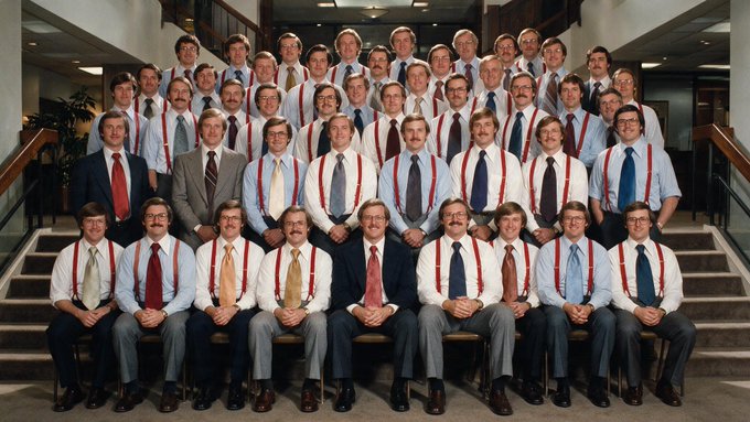 A vintage-style group portrait of approximately 40-50 men wearing matching white shirts, ties, and red suspenders, posed on a grand staircase, resembling a corporate or institutional employee photo.