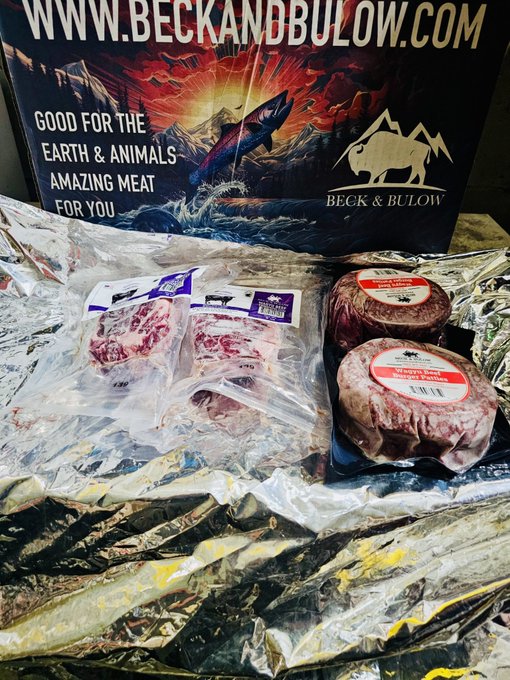 The image features a box of meat, possibly beef or pork, with two packages inside. One package has a red label that reads "Good for the Earth & Animals", while the other has a white label with black text that says "Amazing Meat For You". The background is blurred and appears to be a poster or banner related to Bitcoin.