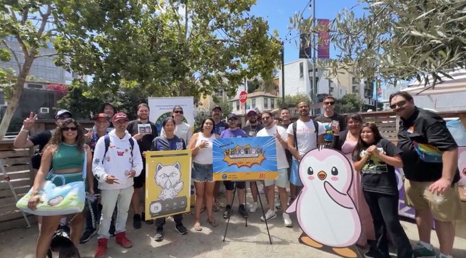 A group of people pose outdoors holding NFT-style cartoon signs, including a cat and a penguin character, with one sign reading 'CREATOR CRYPTO SAFARI'.