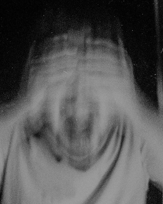 A grainy black and white photograph of a hooded figure with motion blur obscuring facial details, creating an eerie, ghostly appearance.