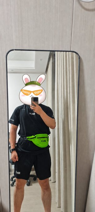 A mirror selfie shows a person wearing black athletic shorts and shirt with a neon green fanny pack, their face obscured by a cartoon rabbit sticker wearing sunglasses.