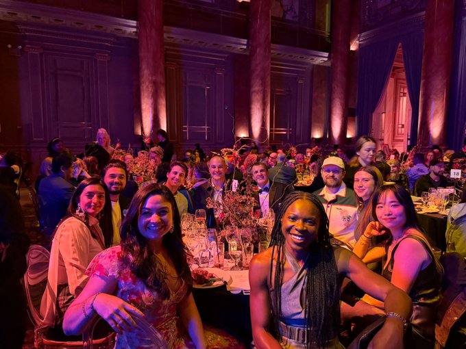 A team of smiling guests poses for a photo at a table during Rhizome's 30th Anniversary Benefit dinner in NYC, with elegant floral centerpieces and purple uplighting on ornate columns in the background. The image captures the celebratory atmosphere of the event where attendees gathered to honor digital art history.