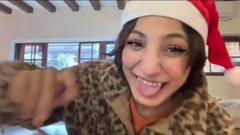 A woman wearing a Santa hat and leopard print jacket smiles and points at the camera in a cheerful pose.