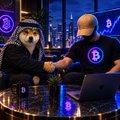 A pixelated cartoon image shows a dog character and a man with a glowing head sitting together at a table with a laptop, with purple Bitcoin logos around them.