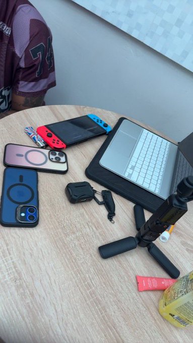 A wooden table displaying various tech accessories, including a Nintendo Switch console, a tablet with a keyboard case, two smartphones in cases with MagSafe rings, earbuds, and a small tripod, with a person in a jersey partially visible.