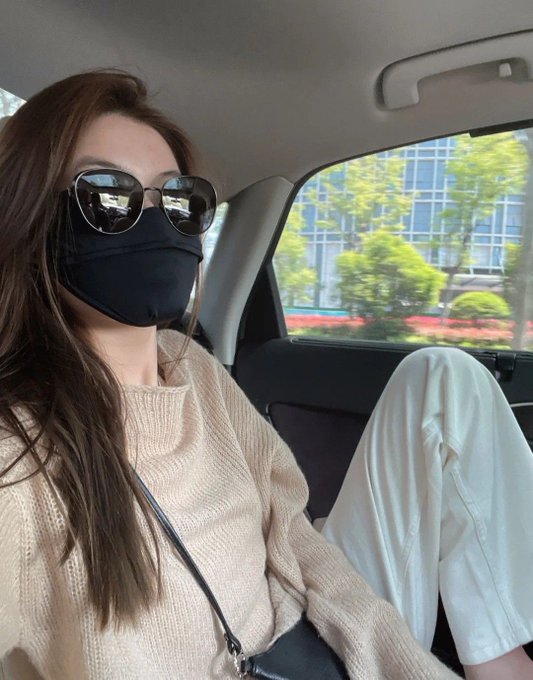 A woman wearing a black face mask and round sunglasses sits in a car wearing a beige sweater, with greenery and a building visible through the window.