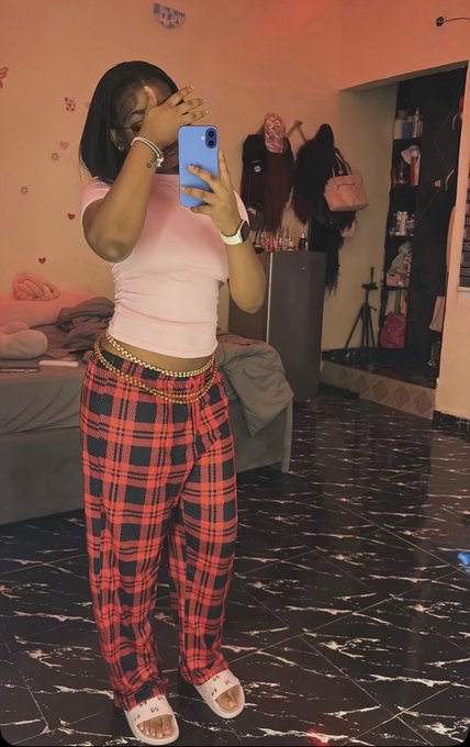A person takes a mirror selfie with a blue smartphone, wearing a pink t-shirt, red and black plaid pants, and sandals, standing in a room with marble floors.