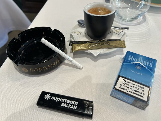 An aerial view of a smoking-related kit on an airplane tray table, including a black Sobranie ashtray, a coffee cup, a Marlboro cigarette pack with Cyrillic text, and a 'Superteam Balkan' lighter.