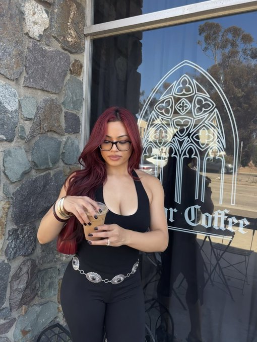 A woman with long red hair, glasses, and a black outfit stands outside a coffee shop holding an iced coffee, with a stained glass window logo visible behind her.