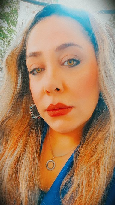 A close-up selfie of a woman with long blonde hair, green eyes, and red lipstick, wearing a blue top with a circular pendant necklace.