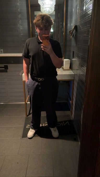 A young person wearing a black polo shirt and pants takes a mirror selfie with an orange phone in a dimly lit bathroom, standing on a "NICE H..." mat, aligning with the "Quant Kid" persona who launched the Gen Z Quant (QUANT) token on Pumpfun.