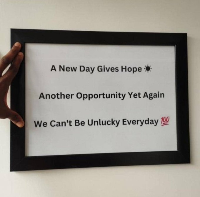 A framed poster with motivational text reading "A New Day Gives Hope, Another Opportunity Yet Again, We Can't Be Unlucky Everyday" with a "100" stamp, held against a white wall by a hand.