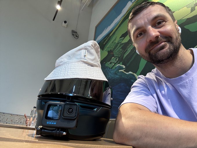 A man with wireless earbuds smiles next to a black motorcycle helmet (with a GoPro attached and a white bucket hat propped on top) sitting on a table, as he recaps a hectic few days filled with World Cup final action, memecoin chaos, Toons minting events, and creator account drama in the Web3 space.