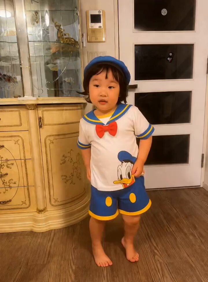 The image features a young child standing barefoot, wearing a Mickey Mouse costume with a hat and shorts. The child appears to be posing for a photo or video, possibly sharing their outfit choice with friends or family members.