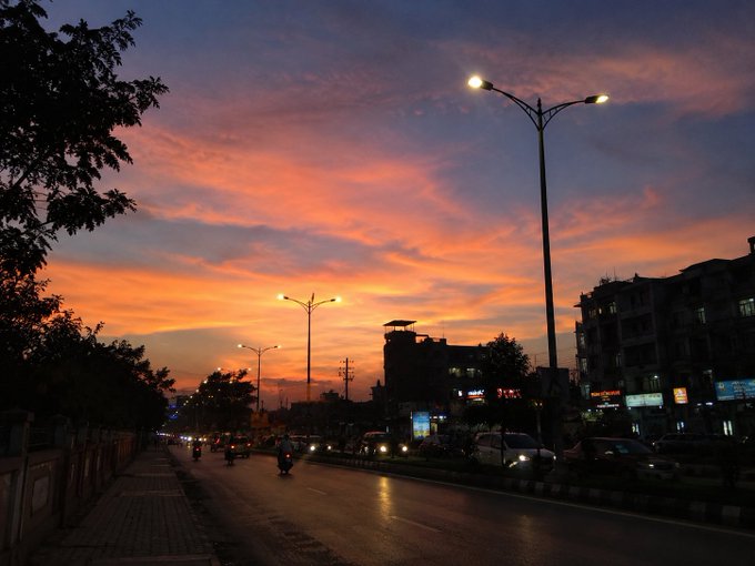 A real photograph of a city street at dusk with a stunning orange and pink sunset sky, illuminated street lamps, silhouetted buildings, and vehicles on the road.