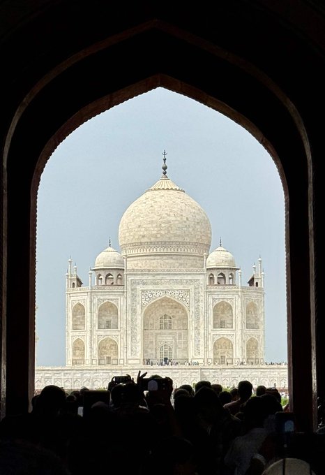 The Taj Mahal viewed through a dark archway silhouette, with crowds of tourists in the foreground taking photos of the iconic white marble mausoleum.
