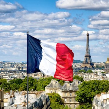 The French tricolor flag waving in the foreground with the Eiffel Tower and Parisian skyline visible behind it under a partly cloudy sky.