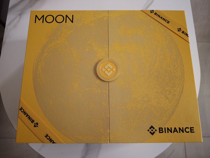 A gold-colored Binance "MOON" gift box or display featuring an image of the moon and Binance logos, tied with a Binance-branded ribbon.