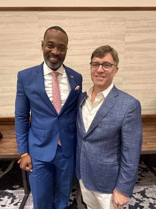 Two smiling men in suits pose together; the man on the left wears a blue suit with a pink polka dot tie, while the man on the right wears a blue blazer and glasses.
