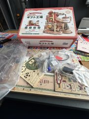 An opened miniature dollhouse kit sits partially assembled, with its "CITY" branded box lid visible in the background and various small furniture pieces, wooden boards, and accessories scattered across the work surface, reflecting the two days of effort required to begin putting it together.