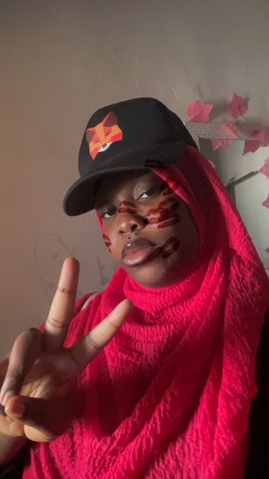 A person wearing a red hijab and a black baseball cap with an orange star logo makes a peace sign while maintaining a serious, unsmiling expression, with red paint or lipstick smeared across their face. The pose and stern look visually reinforce the "Day 1 of not smiling" declaration tied to the $ANSEM bag mentioned in the caption.