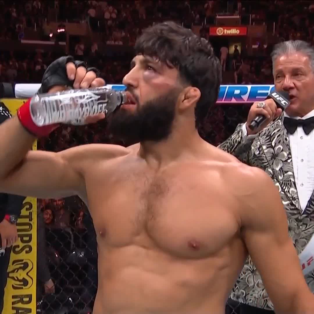 A shirtless bearded UFC fighter, presumably Arman Tsarukyan, hydrates with a water bottle after securing a knockout victory, while a commentator in a floral tuxedo holds a UFC-branded microphone nearby. The background shows a crowded arena with a "twilio" advertisement, fitting the post's celebration of his win at UFC 331.