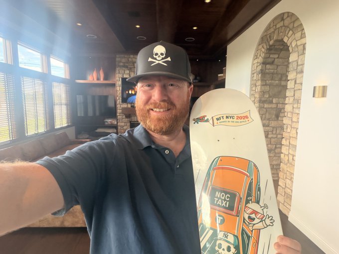 A smiling man wearing a black cap with a white skull logo takes a selfie indoors while holding a skateboard featuring 'NFT NYC 2026' and 'NOC TAXI' graphics.