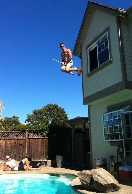 A shirtless man in green shorts is captured mid-air leaping off a second-story roof toward a swimming pool, while two people sit by the poolside watching.