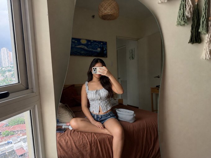 A mirror selfie of a woman with long dark hair sitting cross-legged on a bed, wearing a gray and white striped corset top and denim shorts, with a Starry Night painting visible in the background.